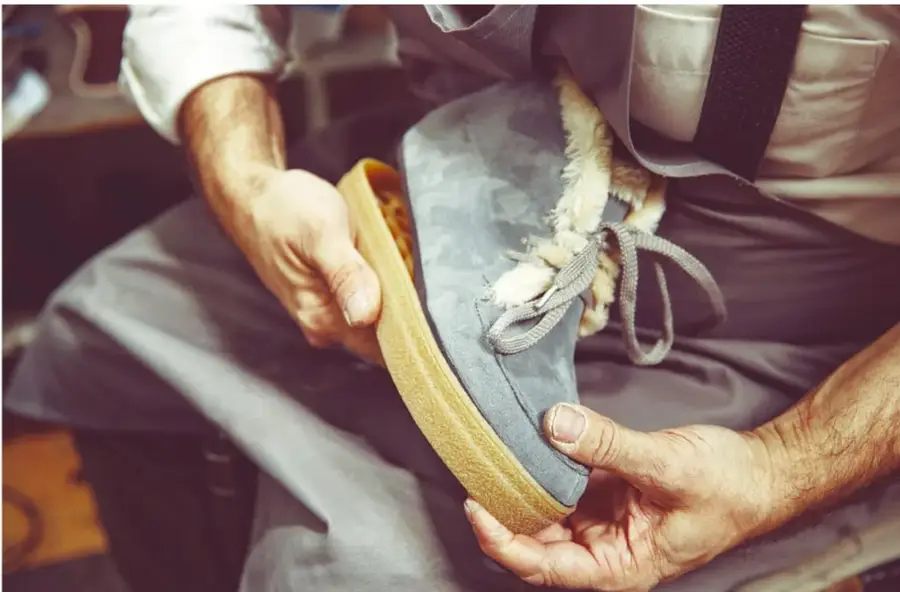 Fotografía de las manos de un zapatero reparando un tenis de piel gris con forro de borrego: sostiene la suela de crepé amarillo mientras trabaja, sentado en su taller con mandil.