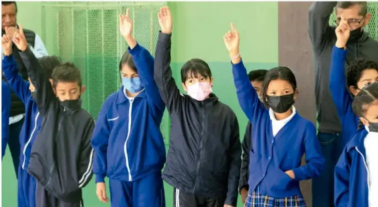 Fotografía de un grupo de estudiantes de primaria con uniforme azul y cubrebocas, de pie frente a una pared verde, con el brazo derecho levantado como señal de participación o compromiso; un adulto los acompaña al fondo.