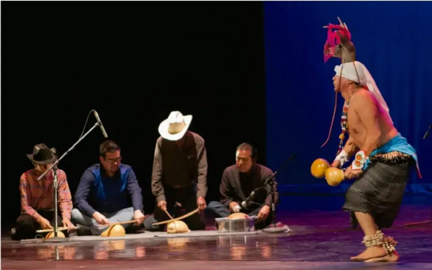 Fotografía de una presentación en escenario: cuatro músicos sentados tocando tambores/cascabeles y un danzante con tocado de venado, piernas con cascabeles, ejecutando la danza.