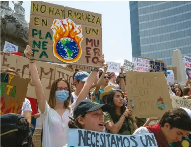 Fotografía a color de una manifestación de jóvenes: sostienen carteles de cartulina, uno con el texto 'NO HAY RIQUEZA EN UN PLANETA MUERTO' con una Tierra en llamas dibujada, y otra pancarta parcialmente visible que dice 'NECESITAMOS UN'.