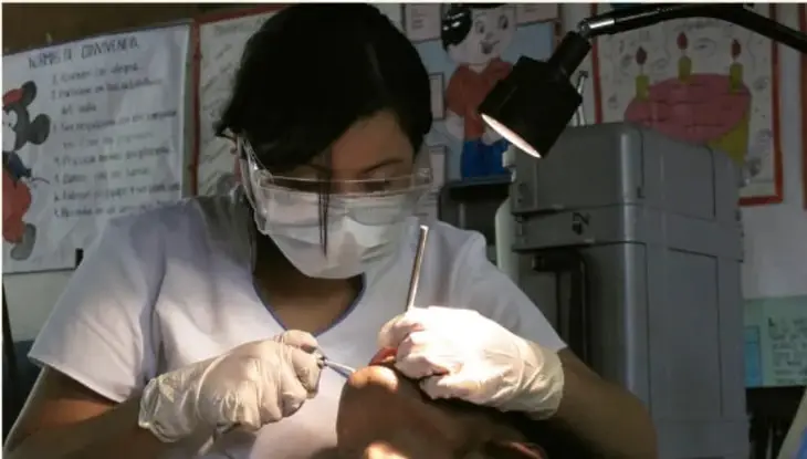 Fotografía de una odontóloga con mascarilla, guantes y lentes protectores atendiendo a un paciente. Al fondo hay carteles con información sobre salud bucal y un equipo dental.