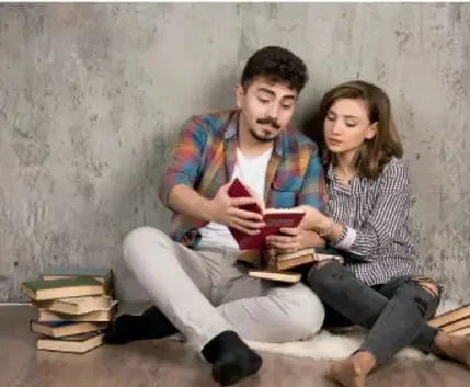 Fotografía de un joven y una joven sentados en el piso junto a una pila de libros, leyendo un libro juntos.