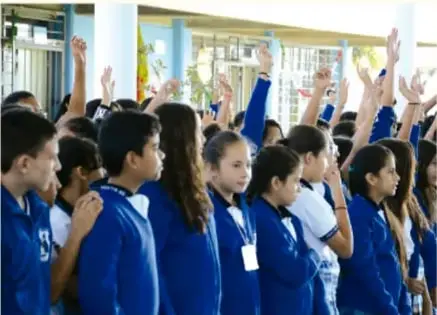 Grupo de estudiantes con uniforme escolar levantando la mano dentro de un aula.