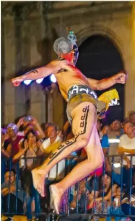 Fotografía de un jugador que aterriza de bruces durante un juego de pelota tradicional, con el público detrás de una reja y un edificio histórico al fondo.