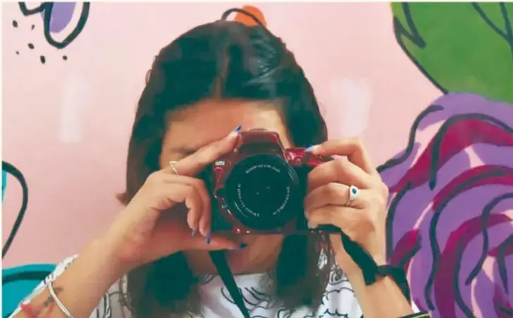 Fotografía de una joven sosteniendo una cámara fotográfica frente a su rostro, con fondo de mural colorido. Pie de foto: Gimenna Schiaffini Rosales, Puebla.