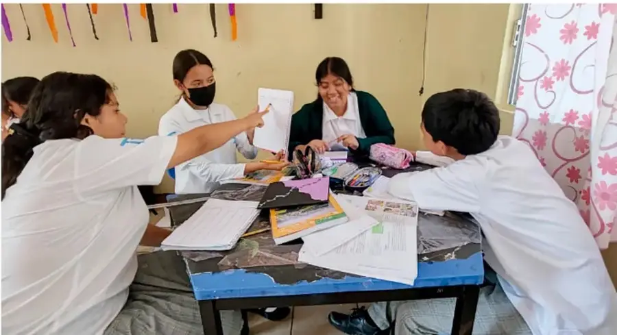 Fotografía de un grupo de estudiantes de telesecundaria con guardapolvos blancos trabajando alrededor de una mesa con papeles, colores y material escolar; una de ellas señala un documento mientras otra lee, en un aula decorada con cintas de colores.