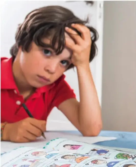 Niño con camisa roja apoyado en una mesa, con la mano en la cabeza, mirando un cuaderno con dibujos. Expresa concentración o preocupación.