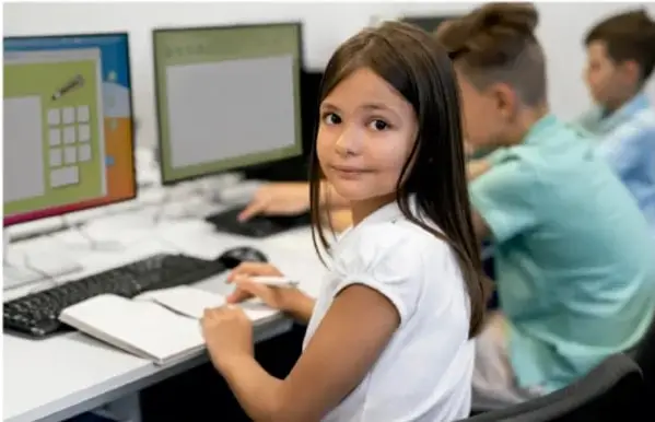 Fotografía de una niña sentada frente a una computadora en un aula, con otros compañeros al fondo, mientras escribe en un cuaderno.