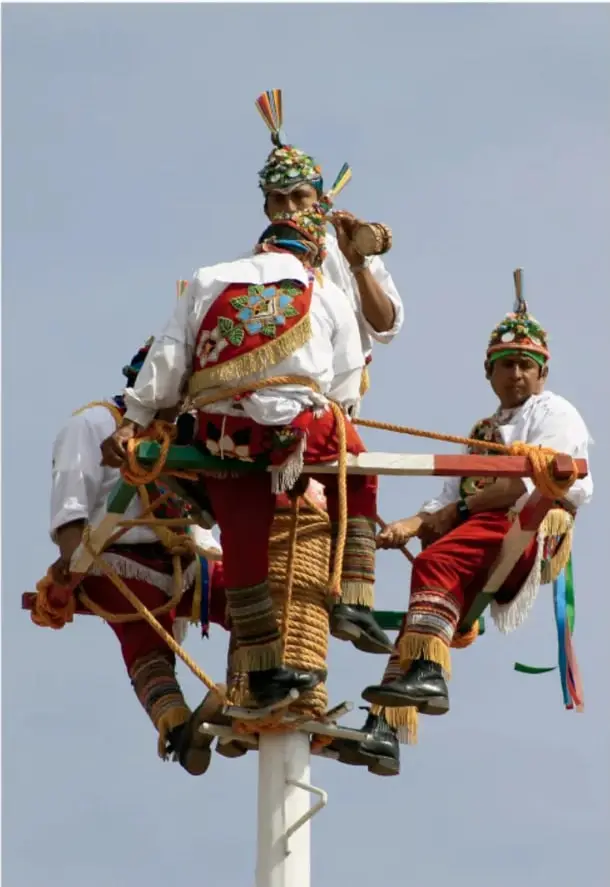 Danzantes ataviados con trajes tradicionales multicolores, sombreros con plumas y botas, amarrados con cuerdas a una plataforma giratoria en lo alto de un palo blanco, practicando una danza ritual comunitaria (posiblemente el Palo Volador).
