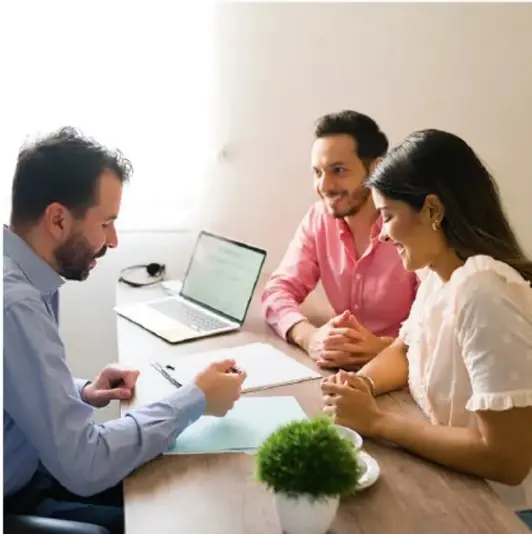 Fotografía de tres personas en una reunión de trabajo alrededor de una mesa con laptop y documentos, representando un entorno de oficina.