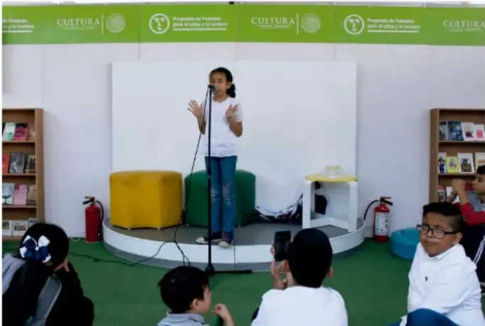 Fotografía de una niña leyendo en voz alta frente a un micrófono en un escenario, con público infantil sentado alrededor.