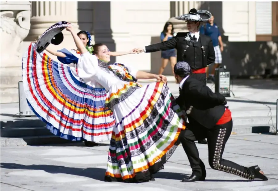 Fotografía de un grupo de danza folclórica mexicana: dos bailarinas con vestidos multicolores de vuelo y dos bailarines con traje charro y sombrero, en una plaza con columnas de piedra.