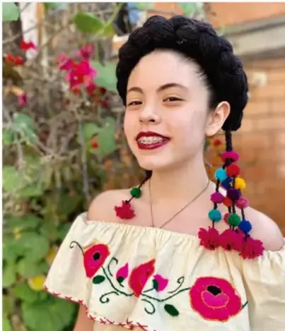 Fotografía de una joven sonriente con vestimenta tradicional mexicana: blusa bordada de flores y trenzas adornadas con borlas y listones de colores (estilo zapoteca del Istmo).