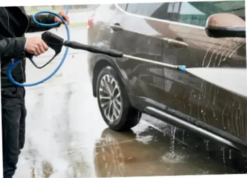 Persona lavando un automóvil con una hidrolavadora; el agua y los residuos escurren al pavimento.