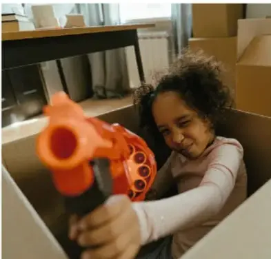 Niña apuntando con una pistola de juguete naranja desde dentro de una caja de cartón, ilustrando el tema de los juegos violentos.