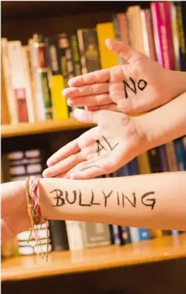 Fotografía de manos con las palmas pintadas con las palabras 'No AL BULLYING' frente a un estante de libros.