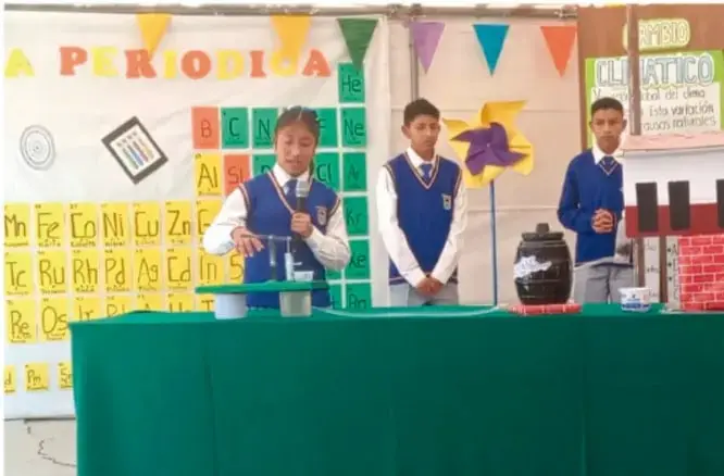 Fotografía de tres estudiantes en uniforme presentando la tabla periódica y materiales sobre una mesa con mantel verde ante su comunidad escolar.