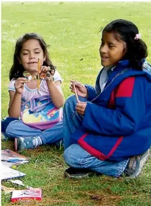 Dos niñas sentadas en el pasto sostienen materiales y dialogan; una lleva chamarra azul con rojo y la otra playera rosa.