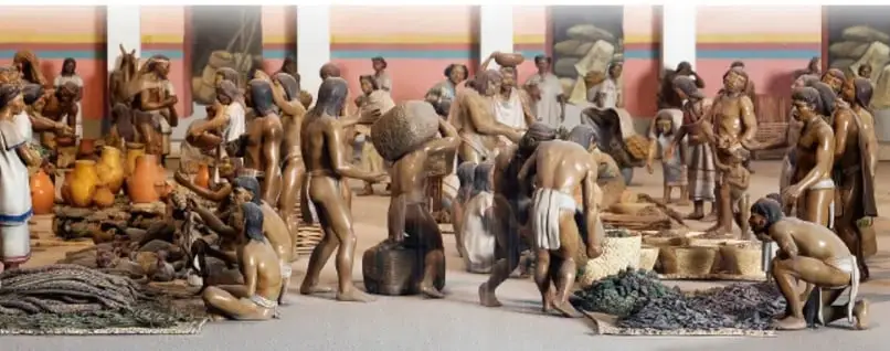 Maqueta o diorama de un mercado mesoamericano con figuras humanas comerciando productos, costales, frutas y distintos objetos.