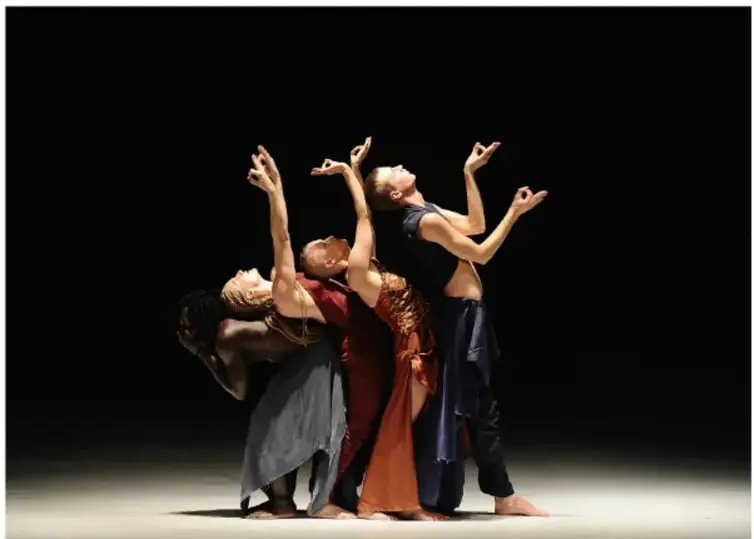 Fotografía escénica de tres bailarines de danza contemporánea con vestuario gris, rojo y azul oscuro sobre fondo negro; corresponde a la obra Aajej de Virpi Pahkinen, fotografiada por Christa Cowrie.
