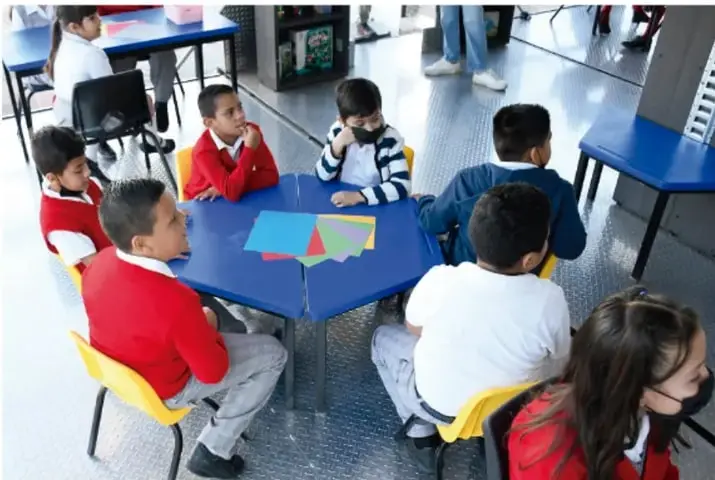 Fotografía de un grupo de estudiantes de primaria sentados alrededor de una mesa hexagonal azul en el aula; algunos miran hacia el centro y otros conversan. Al fondo se ven mesas y sillas escolares.