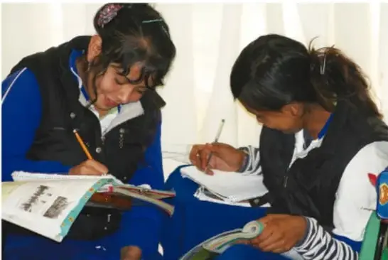Dos estudiantes sentadas en el piso escribiendo en sus cuadernos, colaborando en una actividad de proyecto.