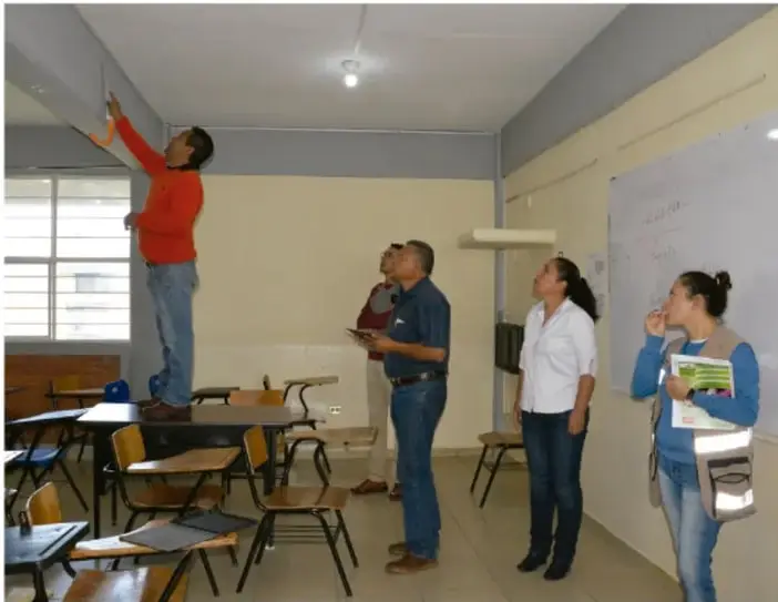 Fotografía de un aula con varias personas: un hombre subido a una silla revisando el techo, y otras personas observando. Representa la revisión de riesgos en la escuela.