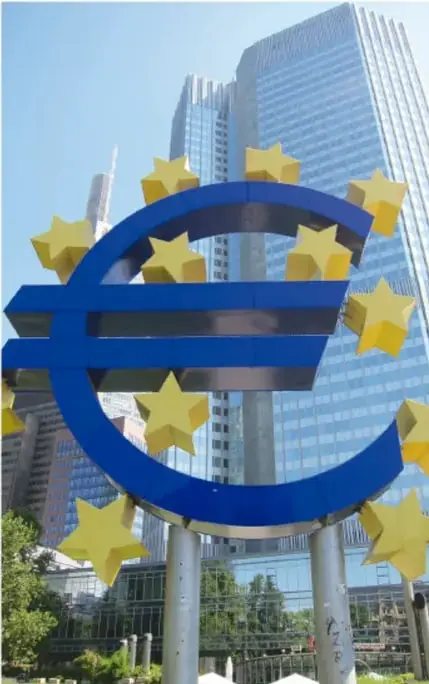 Escultura monumental del símbolo del euro rodeada de estrellas amarillas, frente a edificios modernos de oficinas.