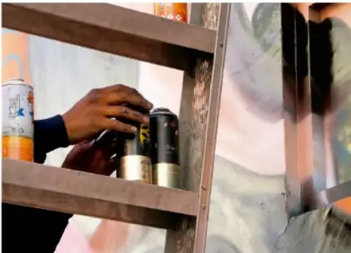 Manos sosteniendo latas de pintura en aerosol frente a un muro con un mural en proceso.