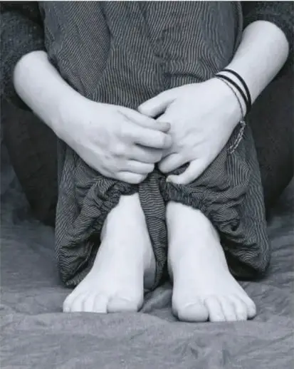 Fotografía en blanco y negro de una persona sentada en el piso, con las manos entrelazadas entre las rodillas y pies descalzos, evocando vulnerabilidad.