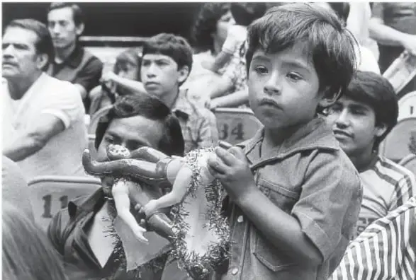 Fotografía en blanco y negro del público en la arena, ca. 1990, con niños y adultos observando el espectáculo, algunos sosteniendo figuras de luchadores.