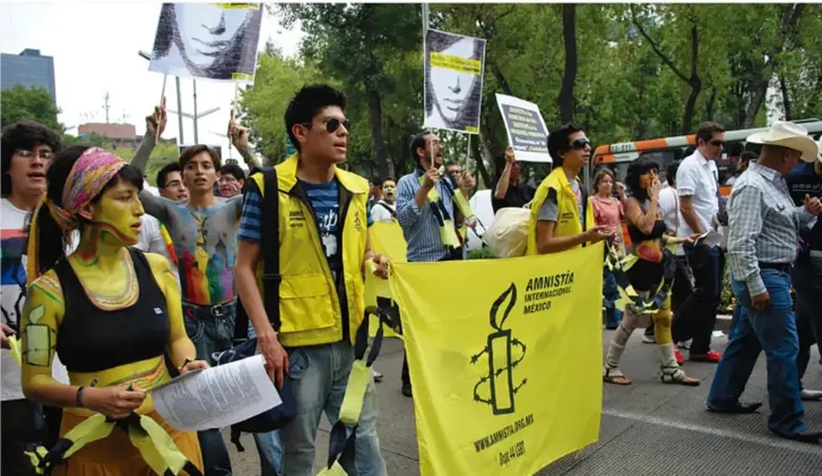 Fotografía de una manifestación callejera: activistas con pintura corporal amarilla y chalecos amarillos desfilan con una manta de Amnistía Internacional México (con el símbolo de la vela entre alambre de púas) y carteles con rostros; al fondo, árboles, un autobús y más participantes.