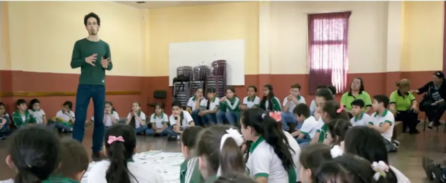 Fotografía de un docente de pie frente a un grupo de estudiantes sentados en el suelo de un salón, durante una actividad escolar.