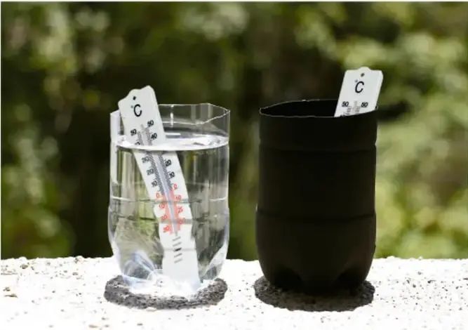 Fotografía de las dos botellas colocadas al aire libre sobre un muro blanco, cada una con un termómetro en su interior; a la izquierda la botella transparente con agua y a la derecha la botella pintada de negro, en un lugar soleado con vegetación de fondo.