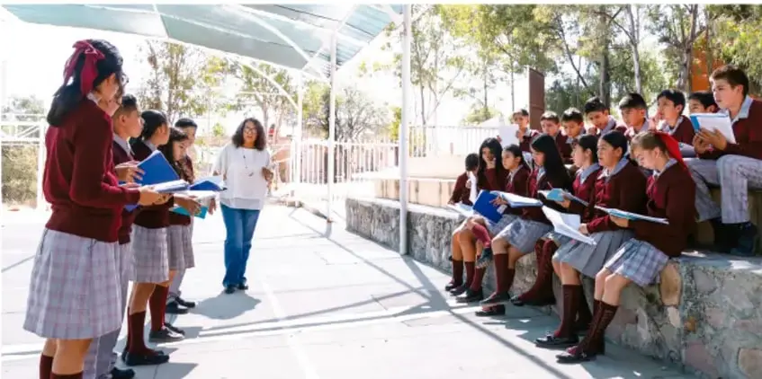 Fotografía de estudiantes de telesecundaria con uniforme, sentados en gradas al aire libre sosteniendo carpetas y escuchando a su maestra durante una actividad de campo.