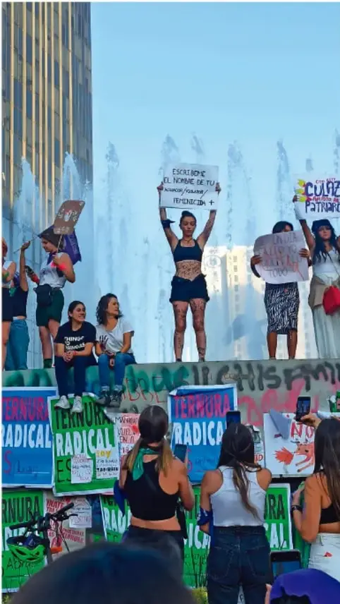 Fotografía de una manifestación pacífica con personas jóvenes sosteniendo carteles con mensajes como 'ESCUCHA EL NOMBRE DE TU HERMANA/COMPAÑERA/AMIGA' y 'TERRURA RADICAL', en un espacio público con fuente.