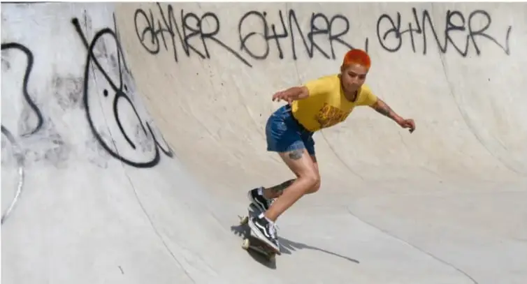 Fotografía de un joven con camiseta amarilla practicando skate sobre una rampa de concreto con grafiti que dice 'OTHER'.