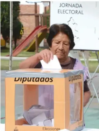 Fotografía de una mujer mayor depositando su voto en una urna transparente marcada con la palabra 'Diputados', en un recinto con el cartel 'JORNADA ELECTORAL'.