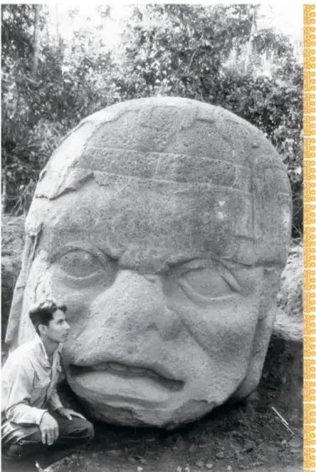 Fotografía en blanco y negro de una cabeza colosal ya desenterrada, con su casco decorado y rasgos serenos, rodeada de vegetación.