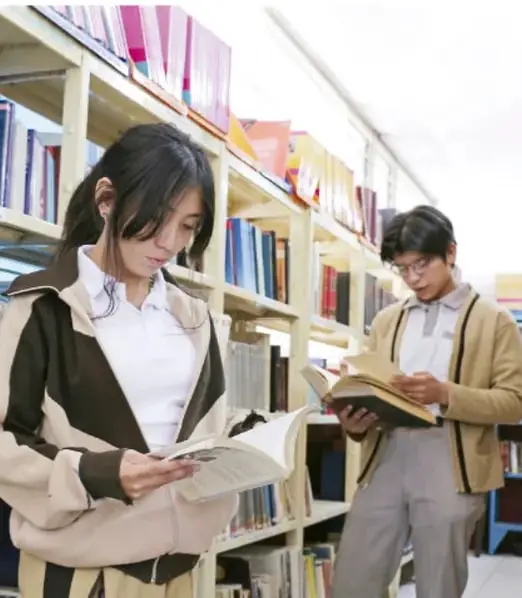 Fotografía de dos estudiantes (una femenina y un masculino) leyendo libros en una biblioteca escolar, entre estanterías con libros.