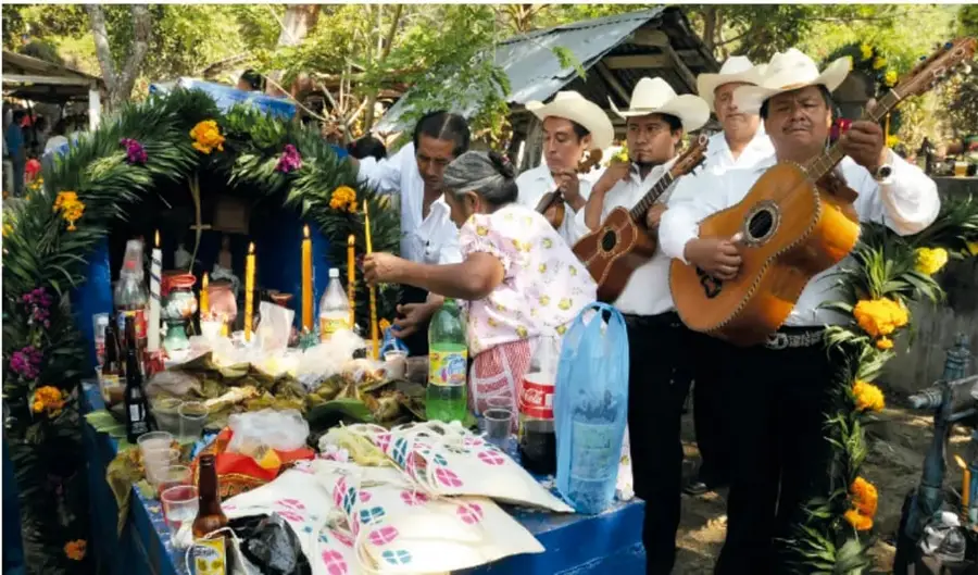 Fotografía de un altar de muertos en Querétaro con flores, velas, botellas y ofrendas, acompañado de músicos con sombreros y guitarras.