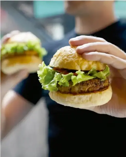 Fotografía de dos personas sosteniendo hamburguesas con lechuga, ilustrando el consumo de comida rápida como ejemplo de riesgo en el ámbito de la salud.