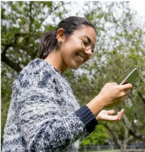 Fotografía de una adolescente sonriente, con lentes, usando su teléfono celular al aire libre, con árboles de fondo.