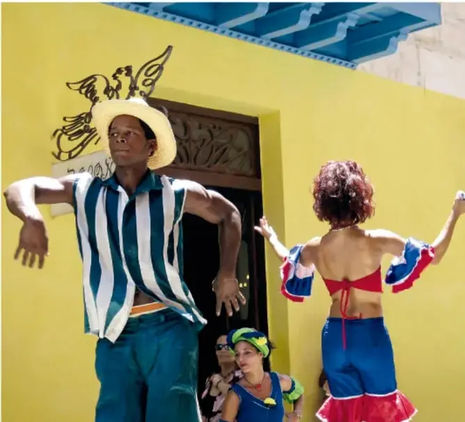 Fotografía de una pareja bailando samba frente a una pared amarilla; el hombre viste camisa a rayas azules y blancas con sombrero, la mujer un top rojo y falda azul con vuelos.