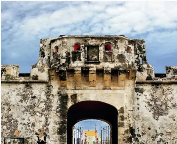 Fotografía de una puerta fortificada de piedra con almenas y pequeños arcos rojos en la parte superior (Puerta de Guadalupe), a través de la cual se ven edificios de colores.