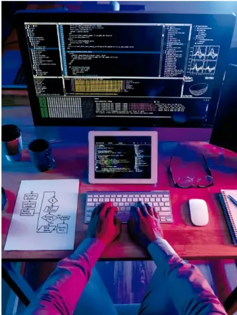 Fotografía de una persona tecleando frente a una computadora con múltiples ventanas de código y gráficas, una tableta y un diagrama de flujo impreso sobre el escritorio; representa el entorno de análisis y programación de datos.