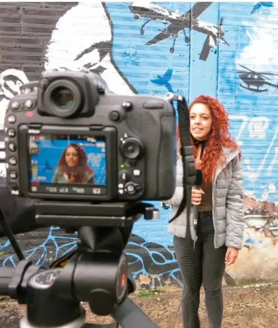 Fotografía de una joven siendo entrevistada frente a un muro con grafiti, con una cámara de video en primer plano registrando la toma.