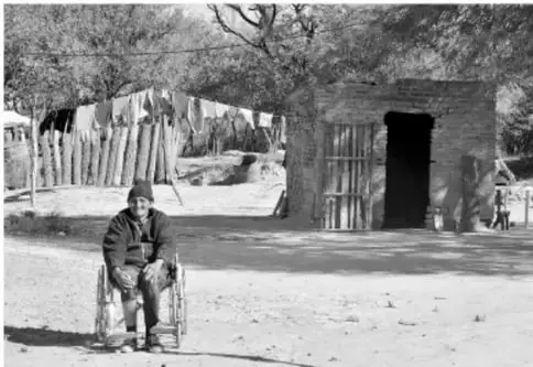 Fotografía en blanco y negro de una persona mayor sentada en una silla de ruedas frente a una vivienda rural de adobe, con cerco de madera, que representa la pobreza y la vulnerabilidad.