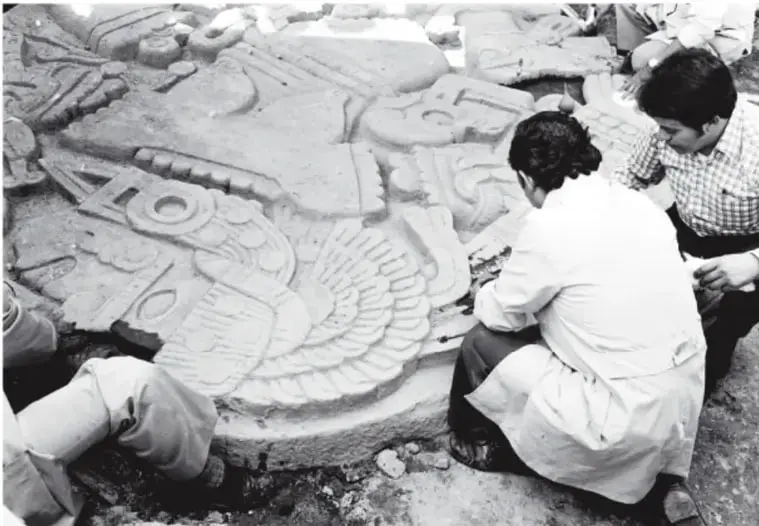 Fotografía en blanco y negro: arqueólogos y trabajadores excavando alrededor de la gran piedra de Coyolxauhqui durante su desentierro; se ve la diosa tallada con su tocado de plumas.