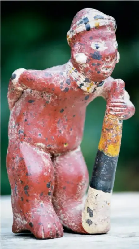Escultura prehispánica de una figura humana de rodillas, policromada en rojo con detalles en amarillo, blanco y negro, sosteniendo un bastón o instrumento multicolor. Representa el arte como medio de expresión cultural.